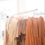Rows of neutral-toned dresses on a clothing rack in a bright boutique with a green plant beside it