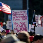 Protest sign with pink handwritten text: 'Don't ever stop trying to make the world a better place. You have POWER. We have POWER' against a crowd backdrop
