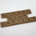 Wooden Scrabble tiles arranged diagonally spell 'CHOOSE YOUR BATTLES' on a white background.